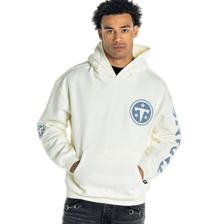 A man wearing a cream-colored Tennessee Titans pullover hoodie with blue graphics, paired with dark gray jeans, against a light gray studio background.