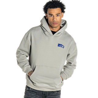 A man wearing a light gray pullover hoodie with a small blue and white Seahawks logo on the left chest, paired with dark gray jeans, against a light gray background.