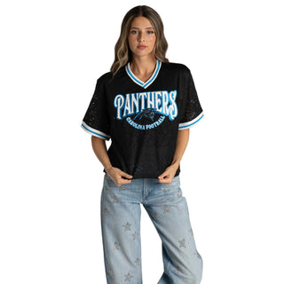 A woman modeling a black short-sleeve V-neck jersey with 'PANTHERS Carolina Football Club' printed in light blue and white on the front, paired with light-wash jeans with star details, against a light gray studio background.