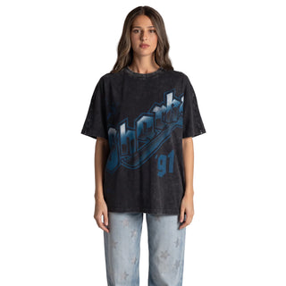 A woman modeling an oversized black acid-wash graphic t-shirt with a blue and white 'Shark' design, paired with light-wash jeans, against a plain light gray background.