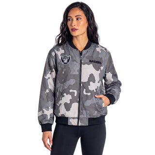 A woman modeling a gray and white camouflage Oakland Raiders bomber jacket against a light gray studio background.